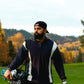 Man with a beard and a black cap holding golf clubs on a golf course with autumn trees in the background