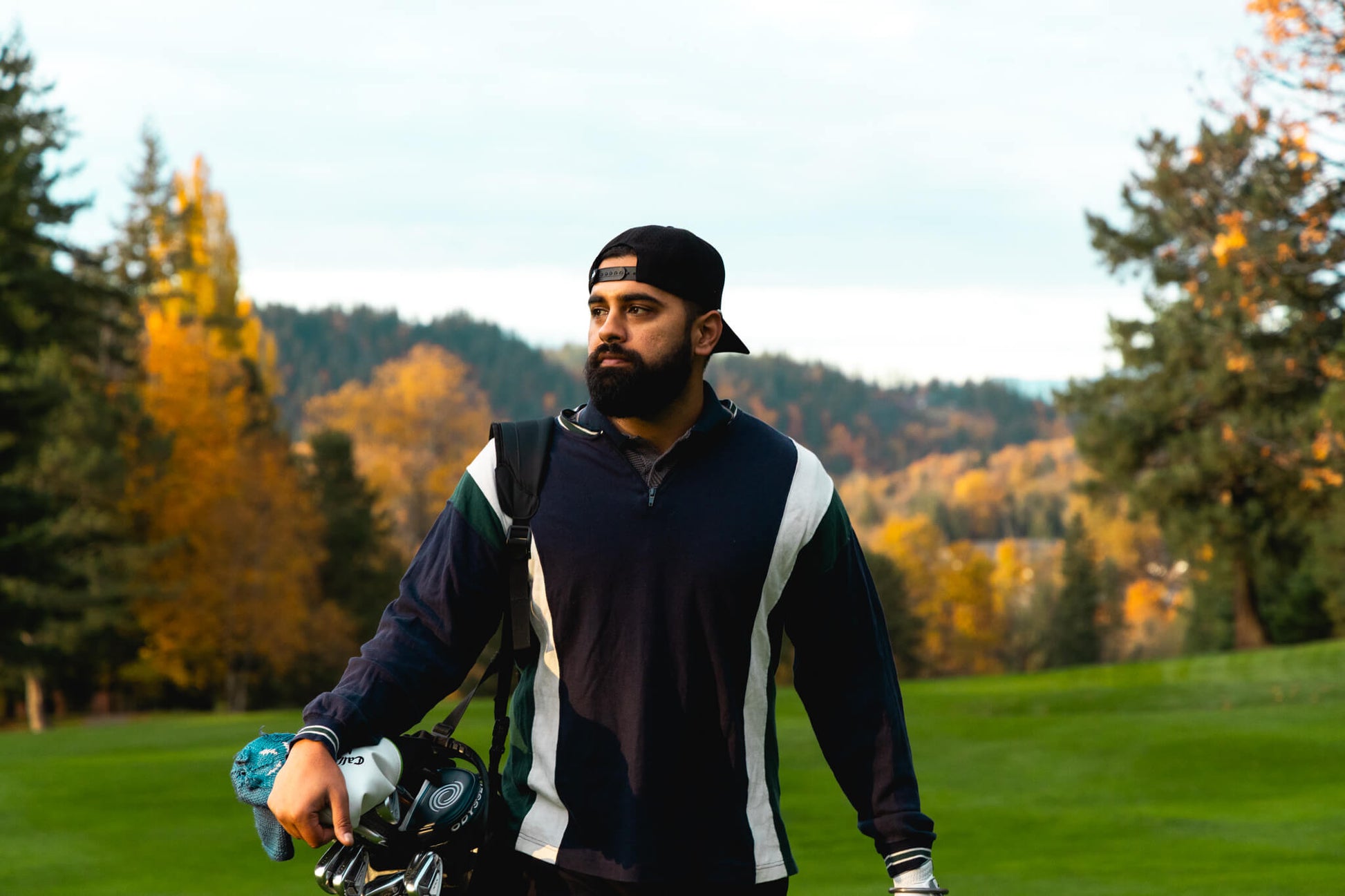 Man with a beard and a black cap holding golf clubs on a golf course with autumn trees in the background