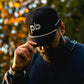 Person wearing a black cap with a Pocket Brush logo, holding the tip of the cap with trees in the background