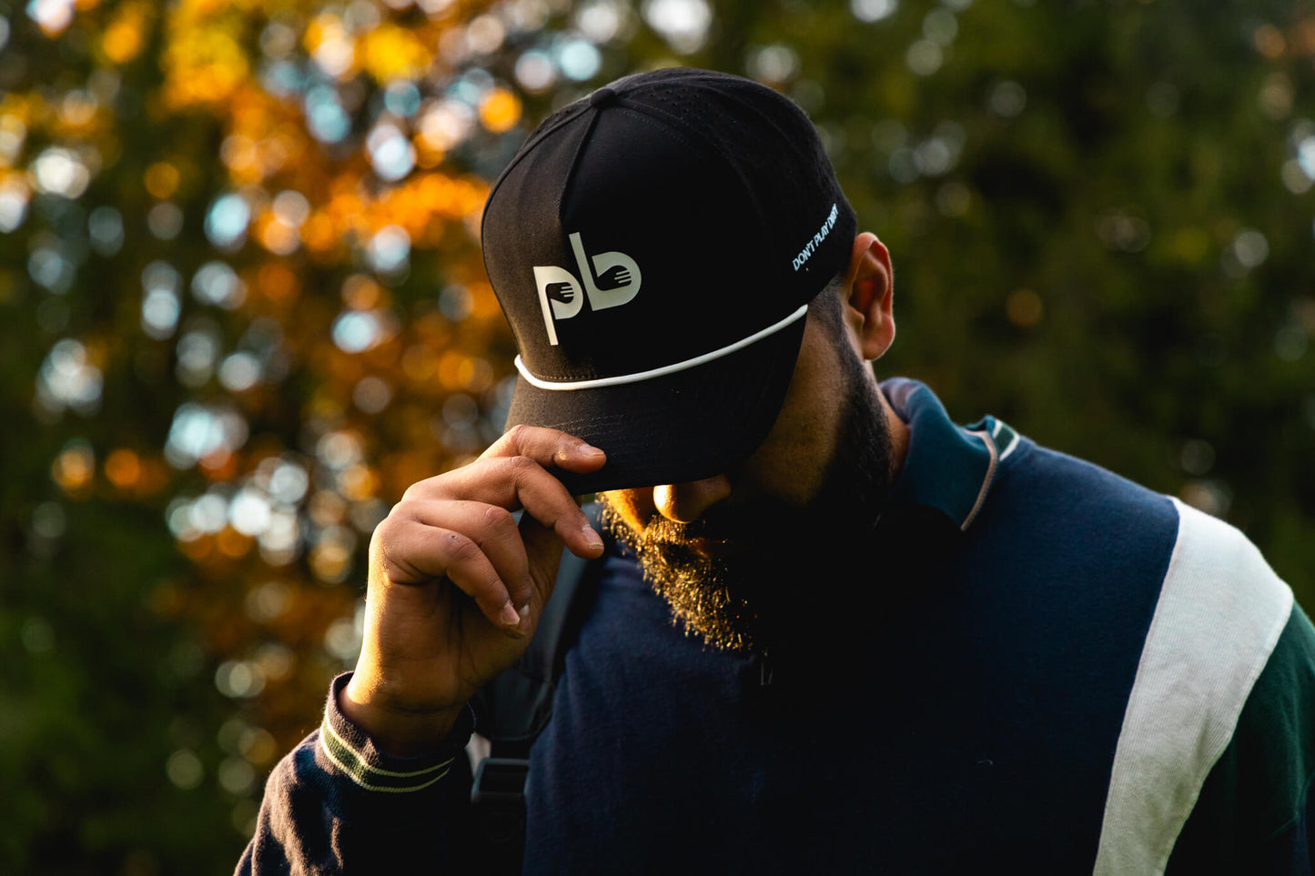 Person wearing a black cap with a Pocket Brush logo, holding the tip of the cap with trees in the background