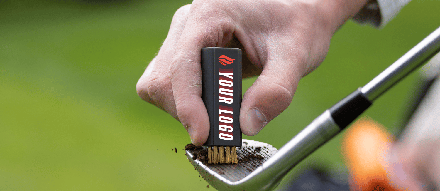 A close-up of a hand holding a small black rectangular cleaning brush with gold bristles over a golf club head, with grass in the background. The brush has a red logo and the text 'YOUR LOGO' printed on it.