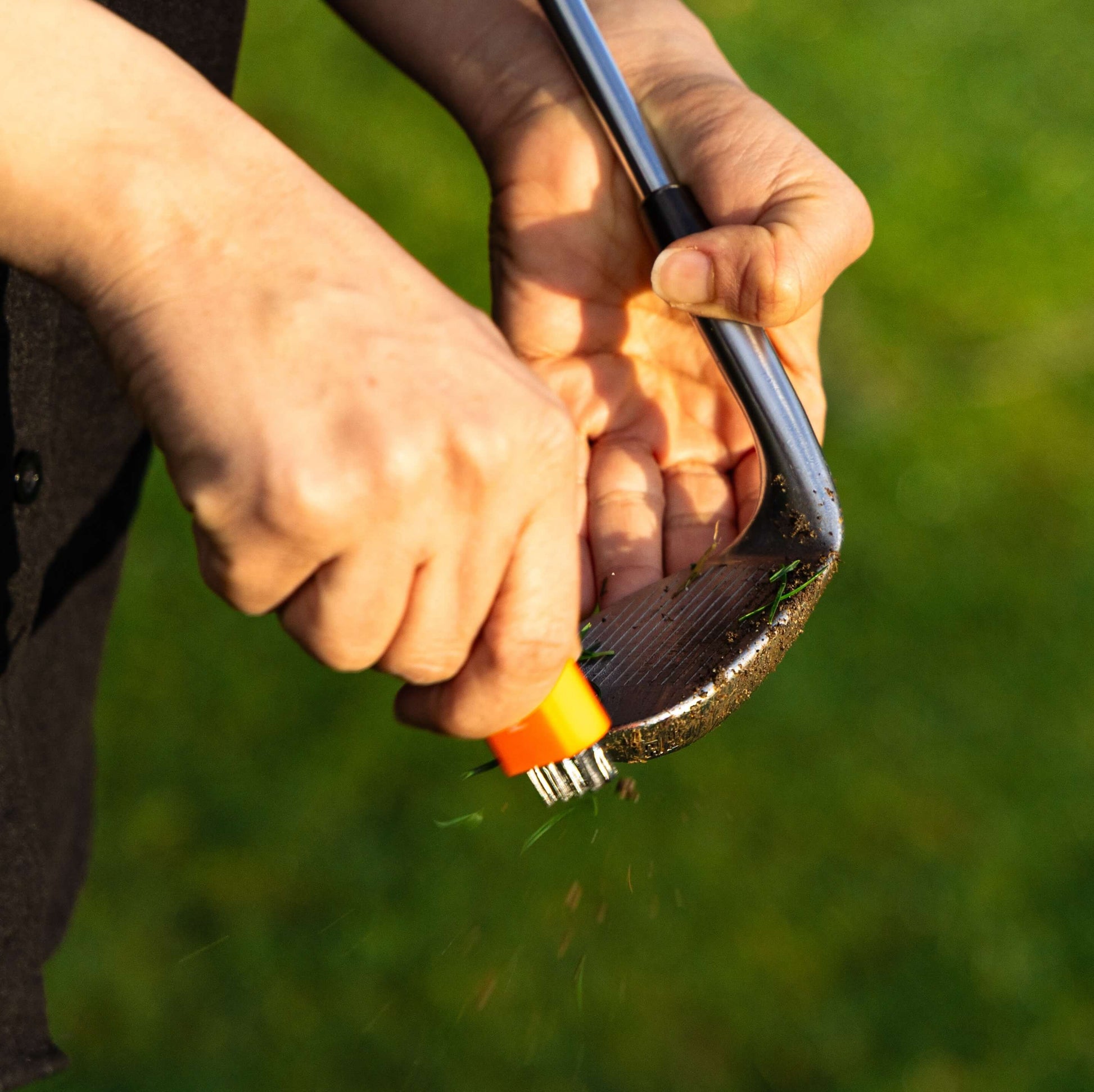 An orange pocket brush is using its brass bristles to wipe the dirt and grass from a dirty golf club face.