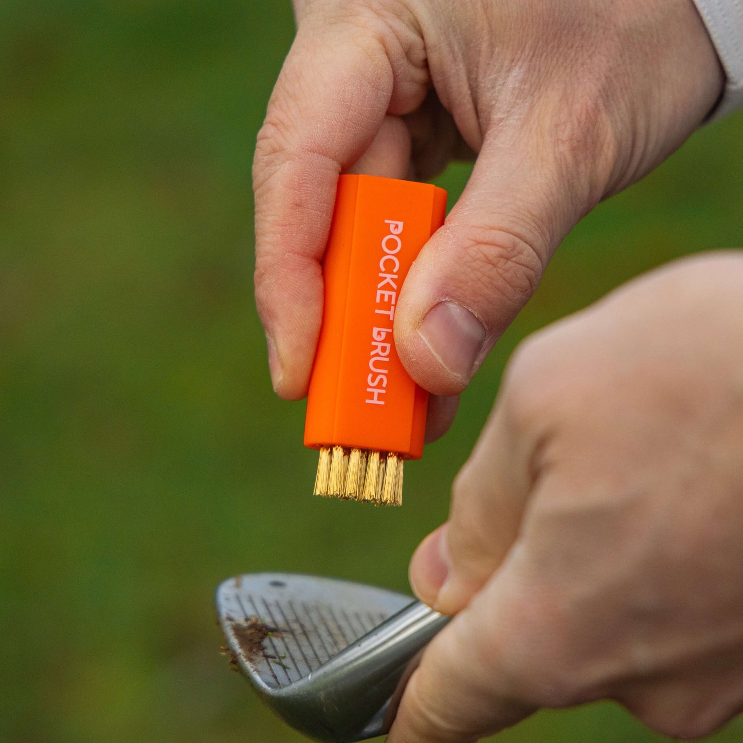 An orange Pocket Brush with its brass bristles exposed is being held above a dirty golf club face.