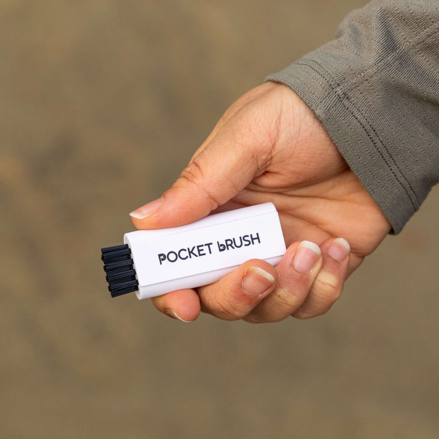 A white Pocket Brush Golf Club Brush and Groove Cleaner is being held with the nylon bristles exposed against a sandy background.