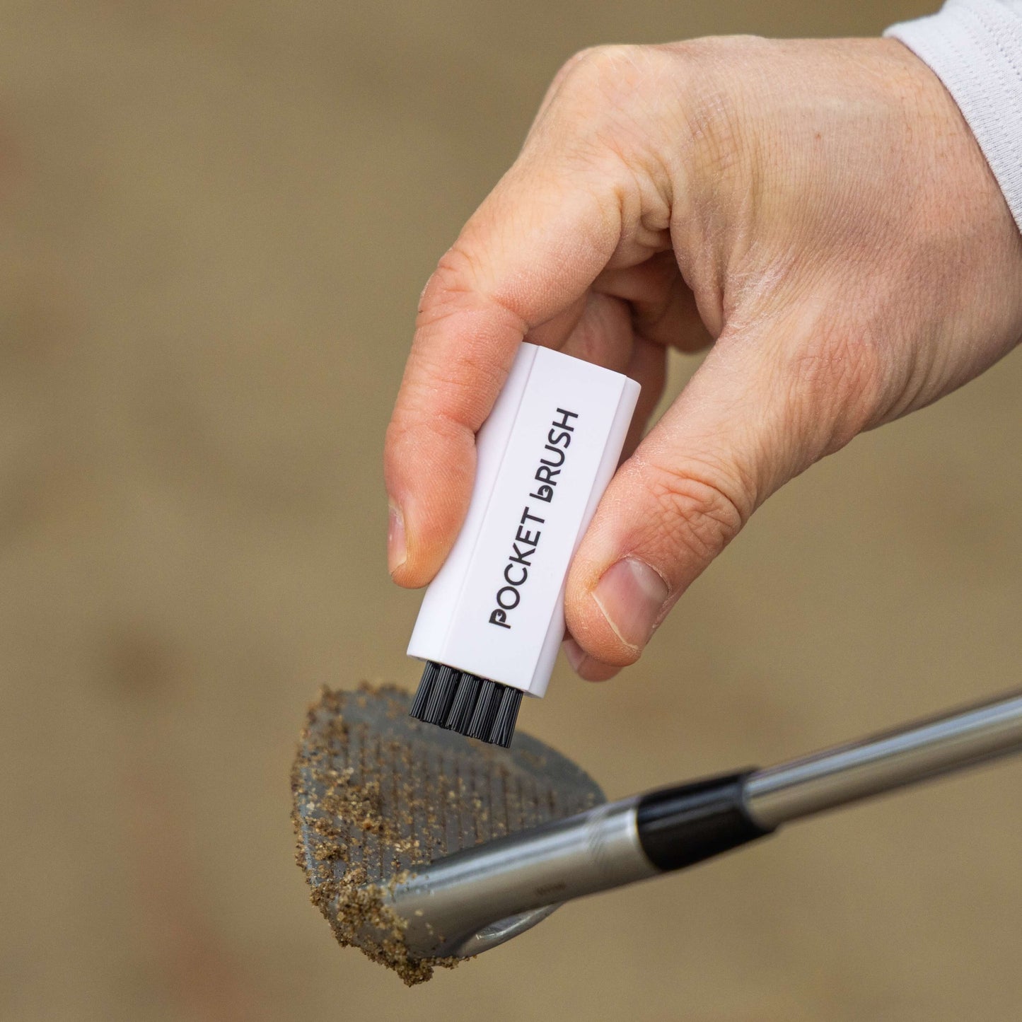 A white Pocket Brush is being held above a sandy golf club face with the nylon bristles exposed.
