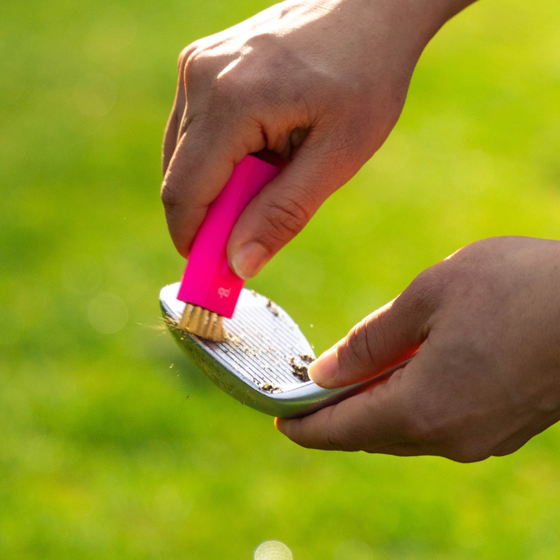 A bright pink Pocket Brush is cleaning the surface of a golf club face using its brass bristles.