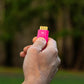 A hand holding a small pink pocket brush with the brass bristles exposed, photographed outdoors against a blurred green and brown natural background.