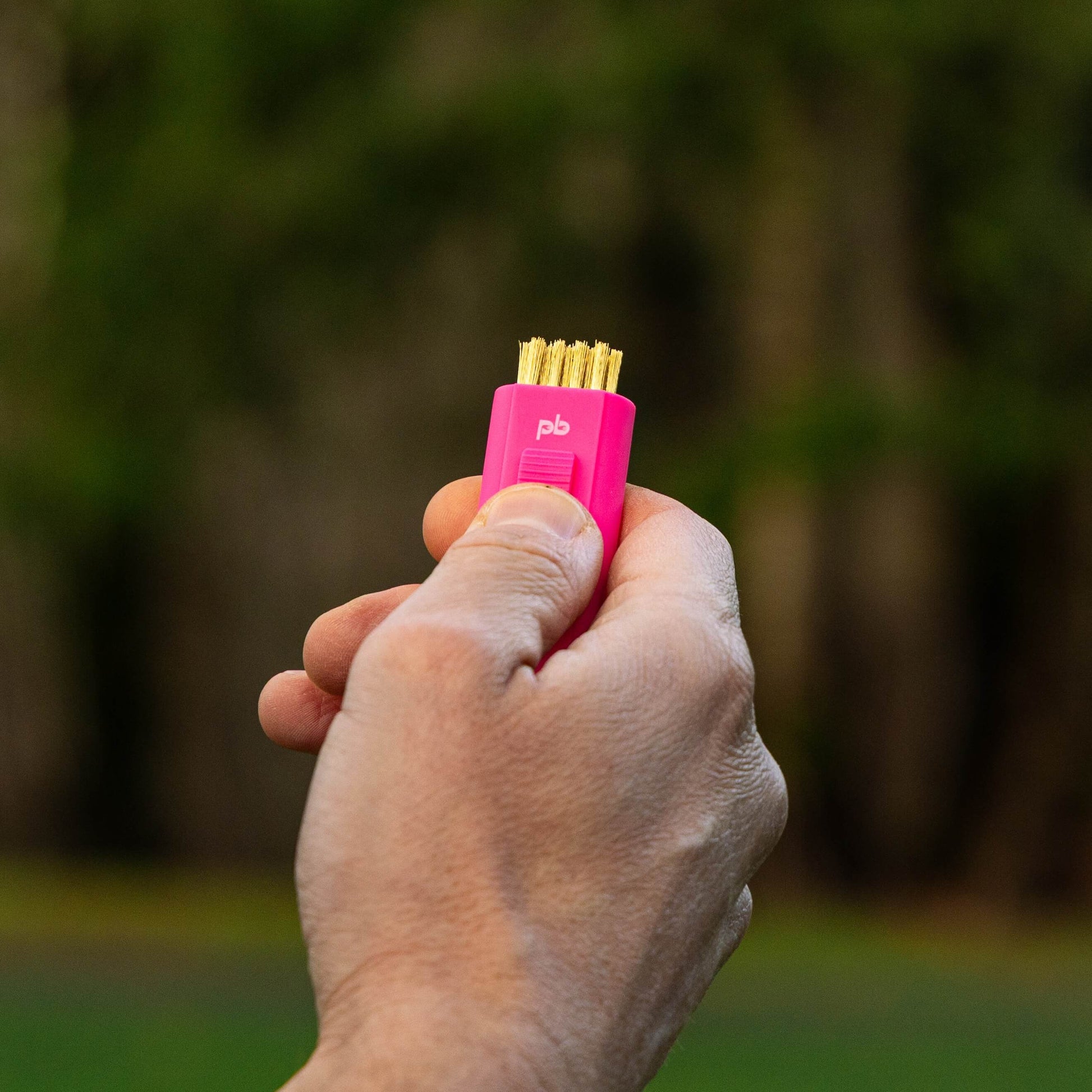 A hand holding a small pink pocket brush with the brass bristles exposed, photographed outdoors against a blurred green and brown natural background.
