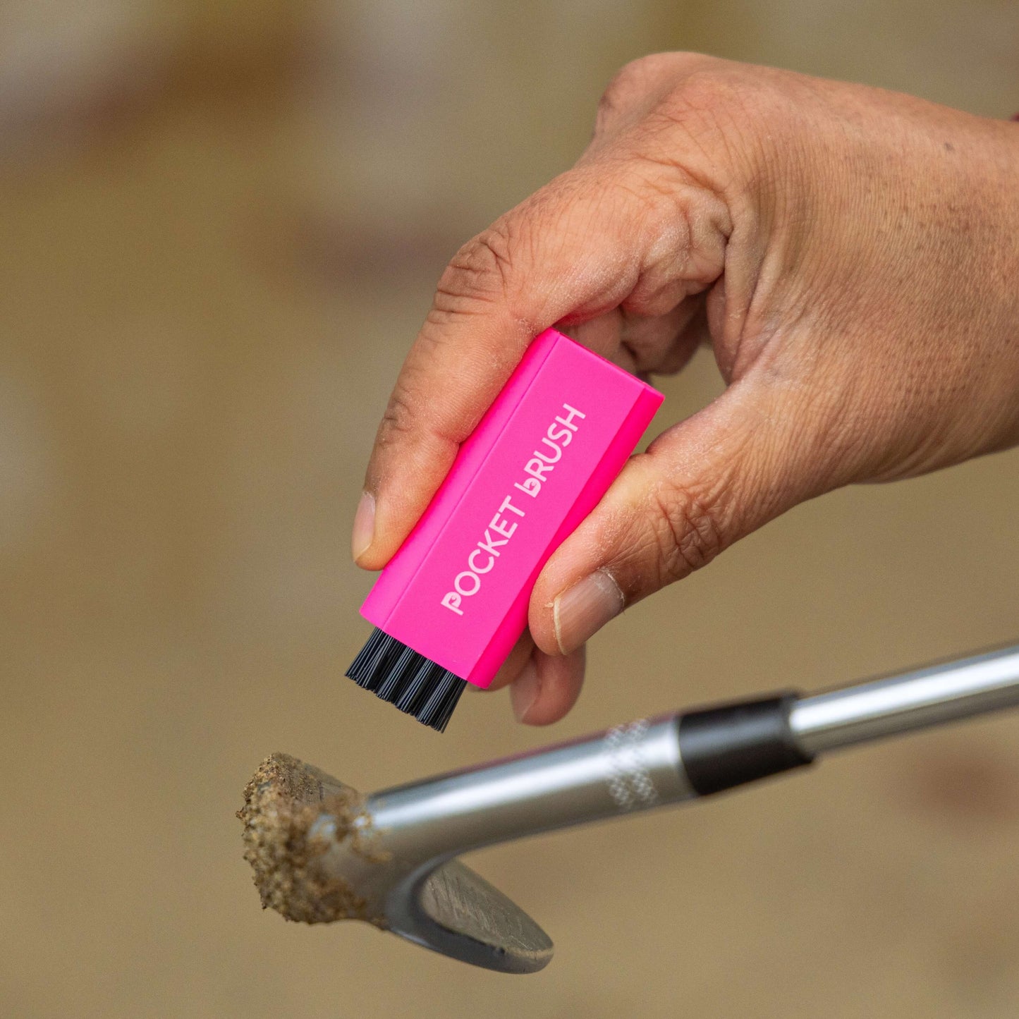 A bright pink Pocket Brush with its nylon bristles exposed is being held overtop of a sand covered golf club.