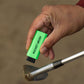 A neon green Pocket brush with its nylon bristles exposed is being held up over a sandy golf club face.