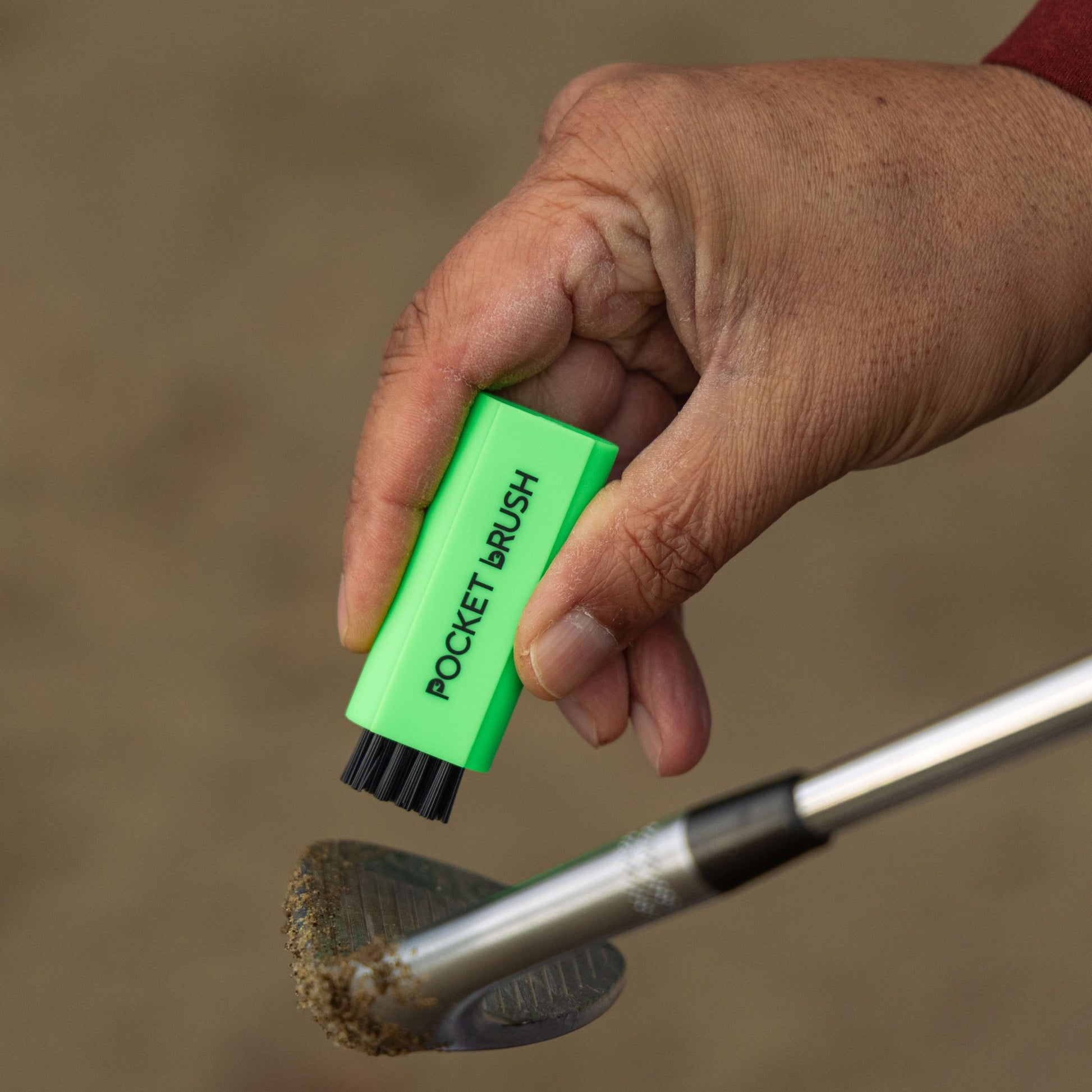 A neon green Pocket brush with its nylon bristles exposed is being held up over a sandy golf club face.