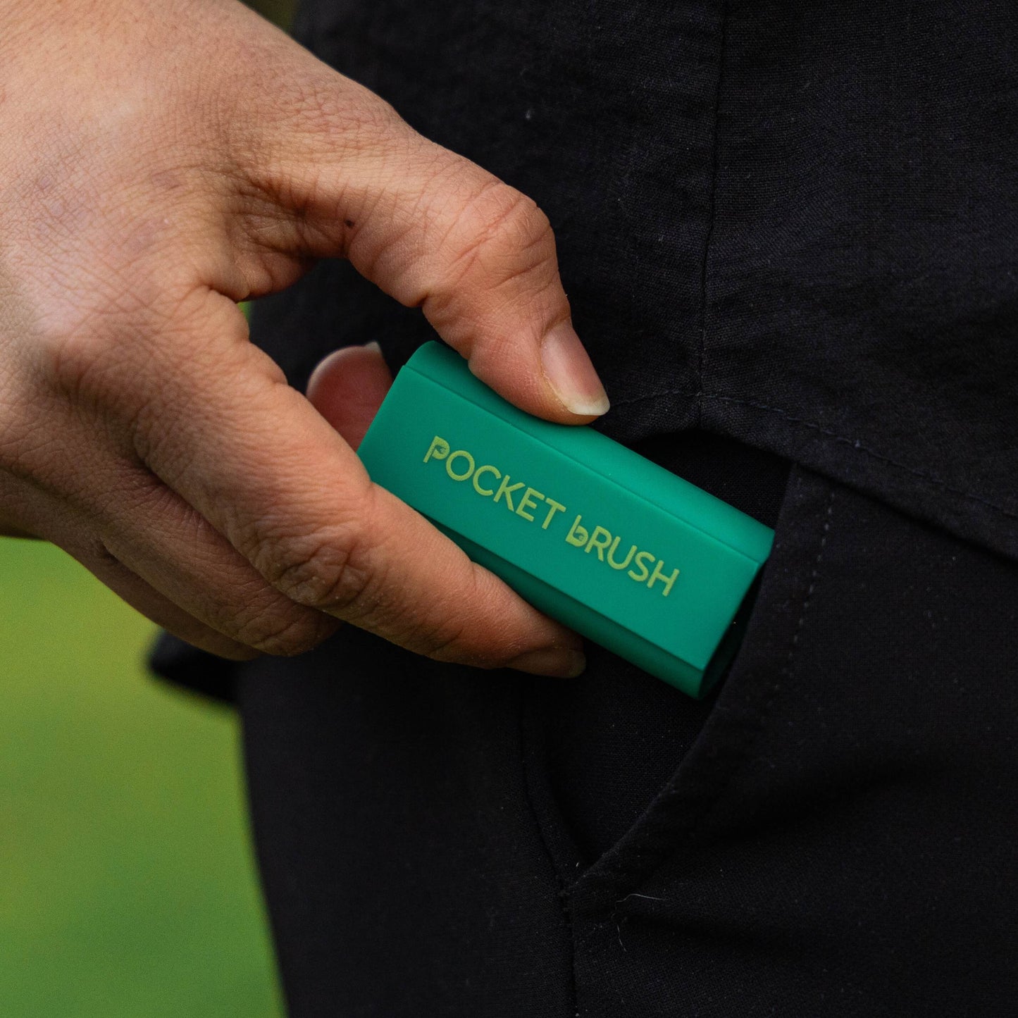 A close-up of a hand sliding a small teal green pocket brush into a black jacket pocket. The pocket brush features gold lettering that reads 'POCKET BRUSH'.