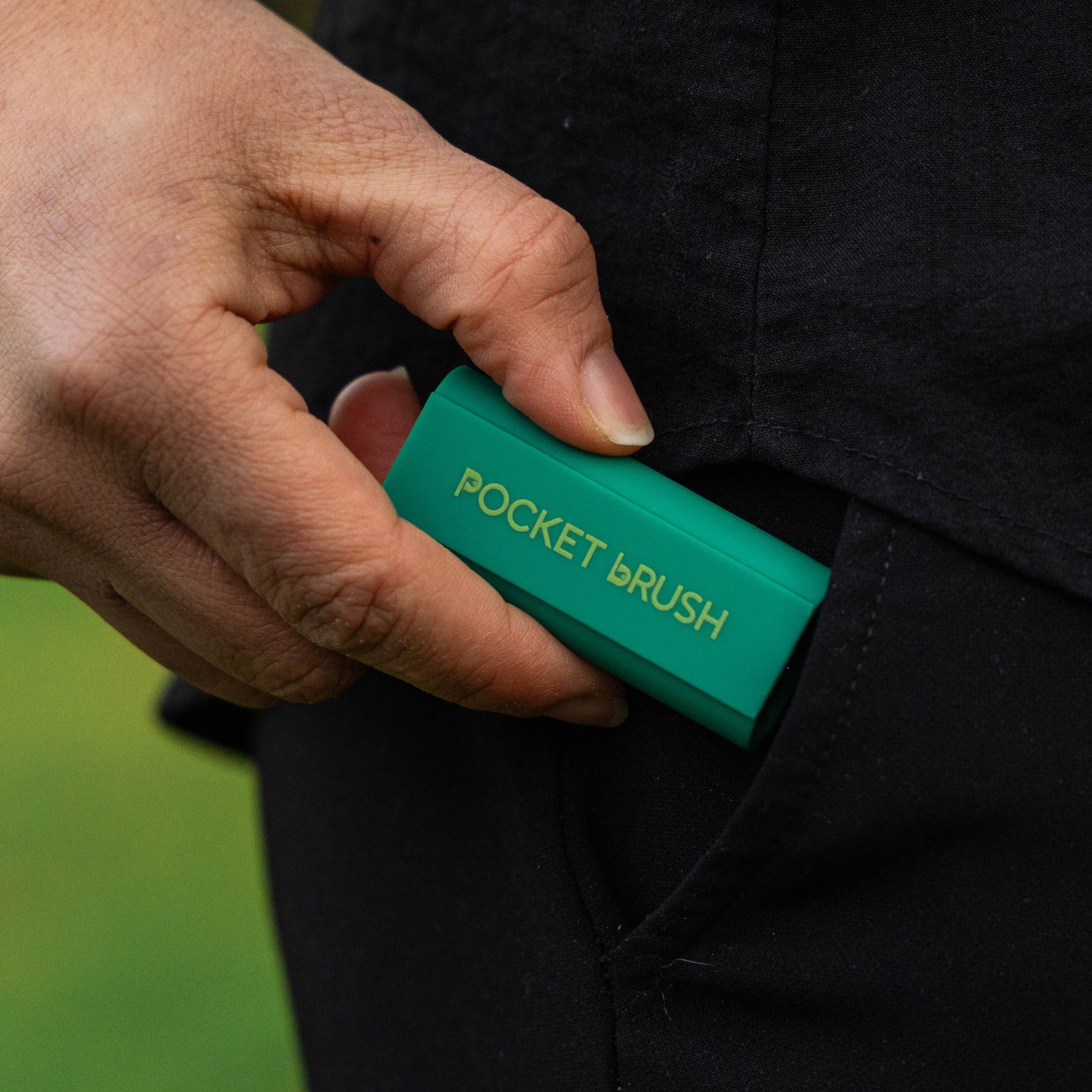 A close-up of a hand sliding a small teal green pocket brush into a black jacket pocket. The pocket brush features gold lettering that reads 'POCKET BRUSH'.
