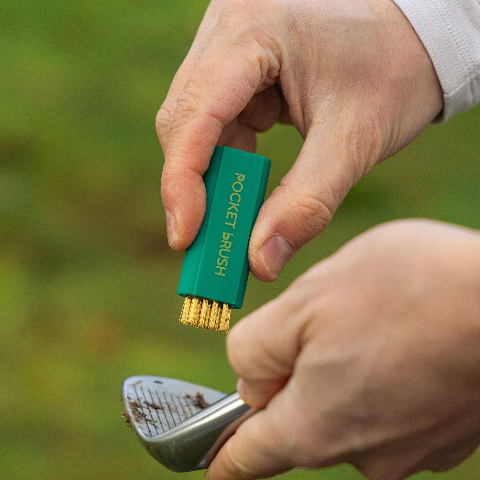 A masters green Pocket Brush is being held with the brass bristles exposed overtop of a dirty golf club face.