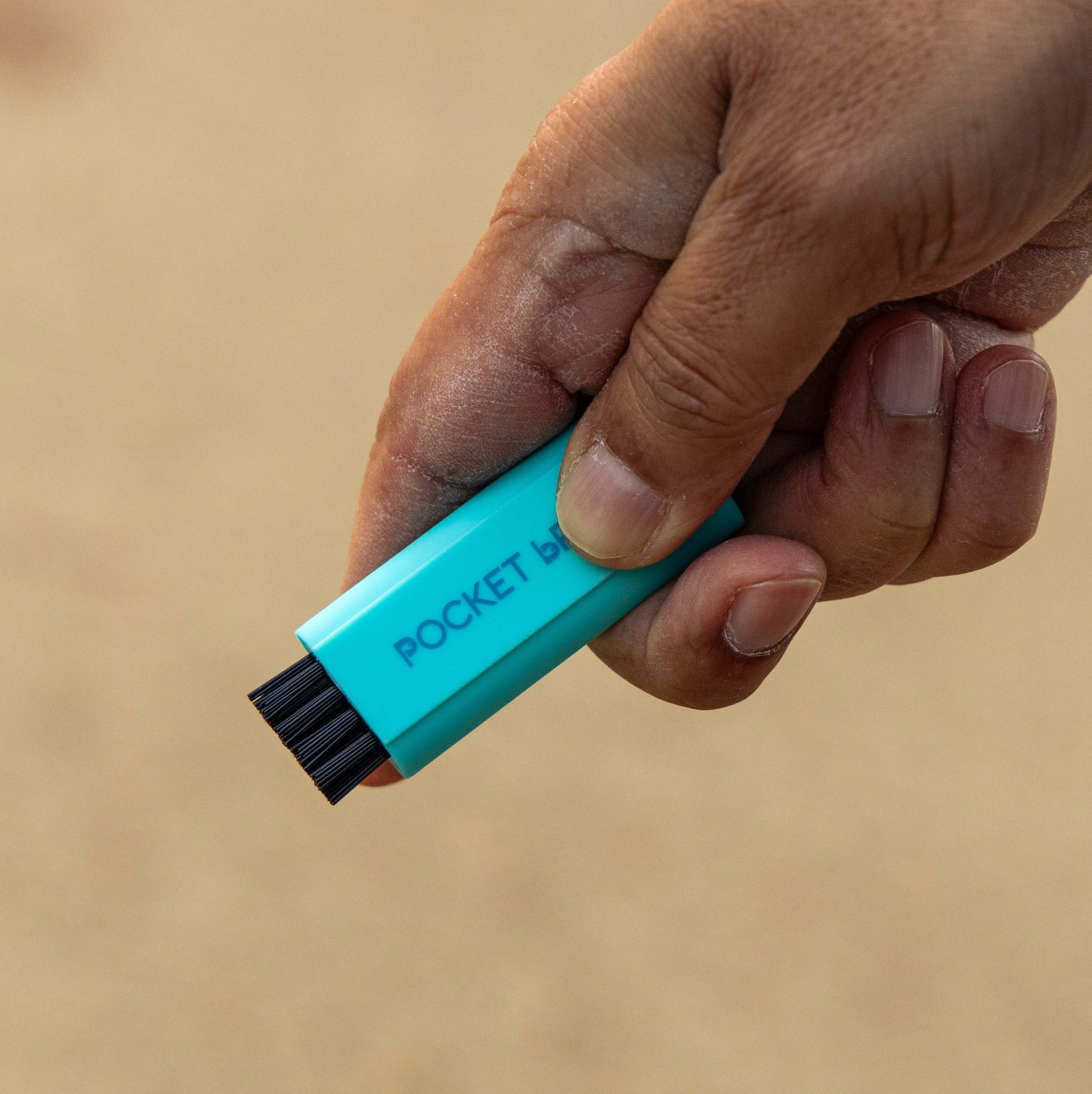 A light blue Pocket Brush is being held up over a sandy background with its nylon bristles exposed