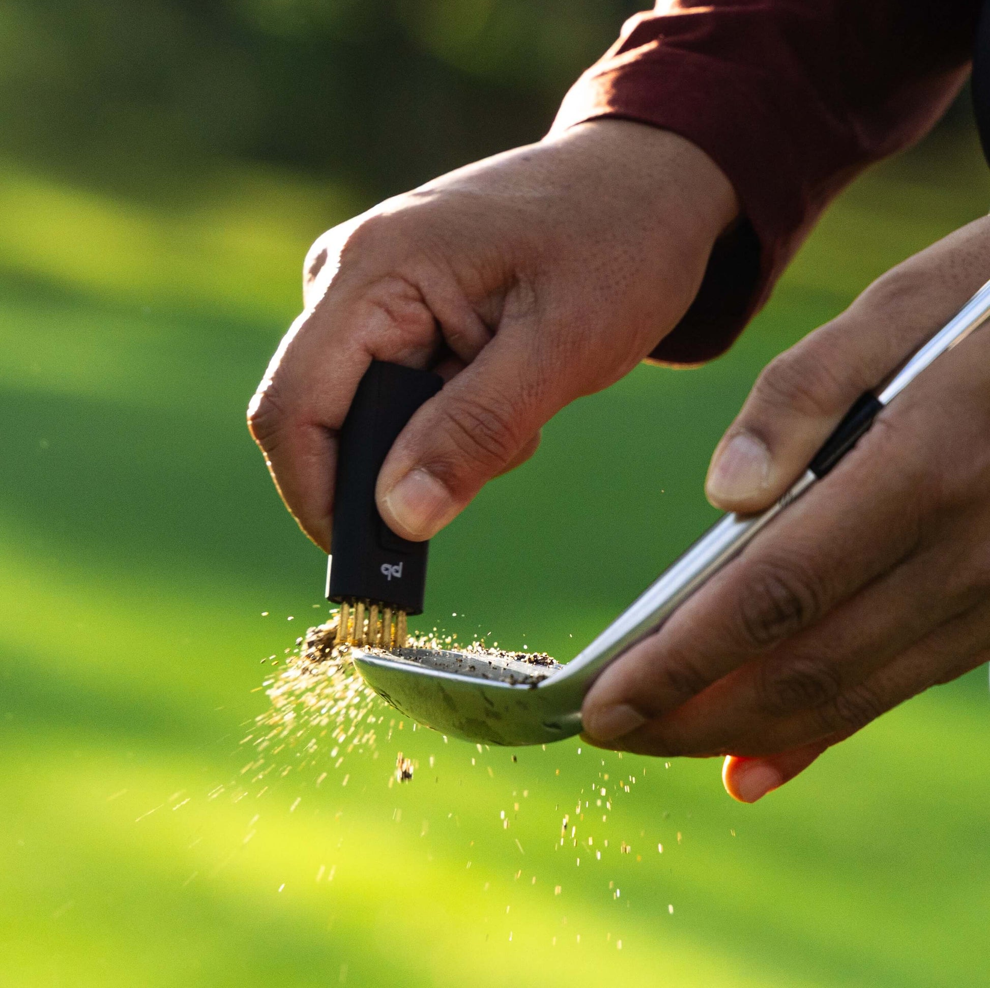 A black Pocket Brush is using its brass bristles to remove the dirt from a dirty golf club face.