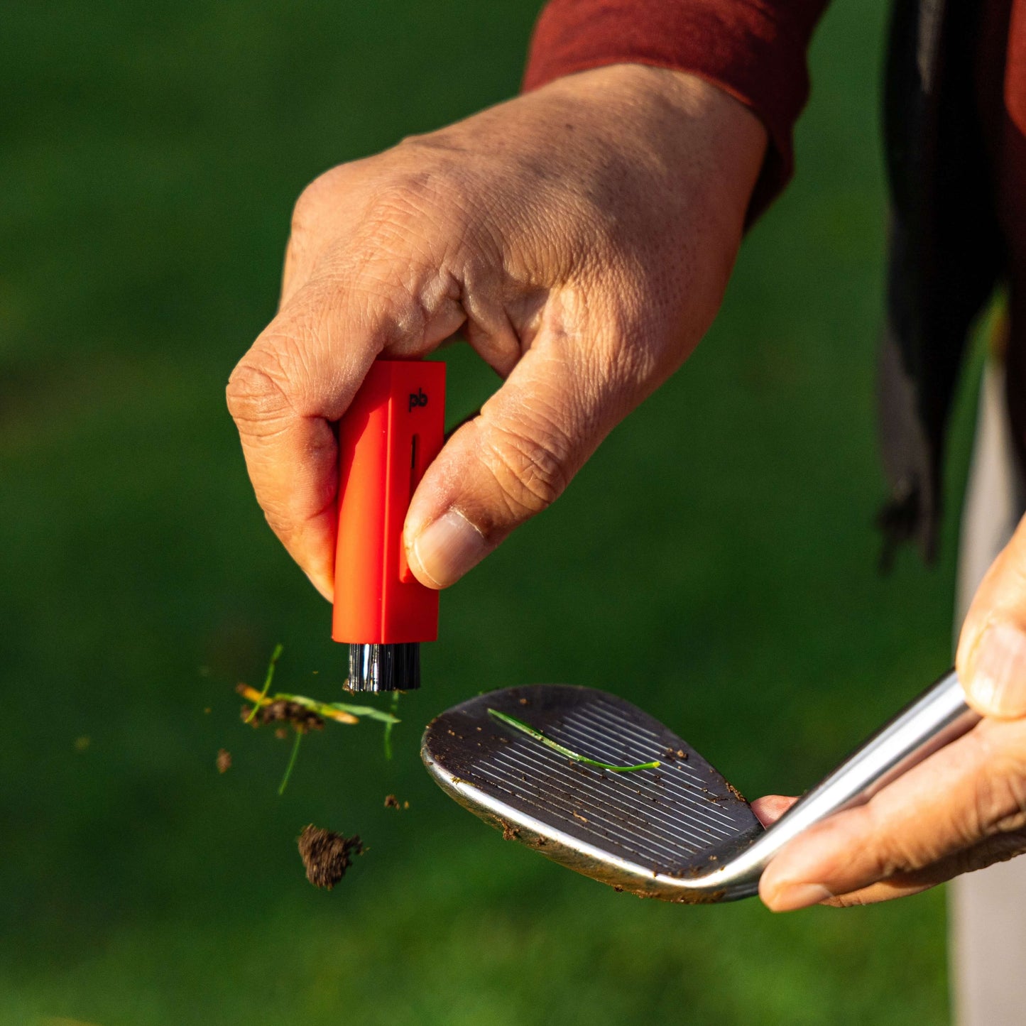 A red Pocket Brush with the nylon bristles exposed is cleaning the face of a dirty golf club. Debris flies towards the left side of the screen