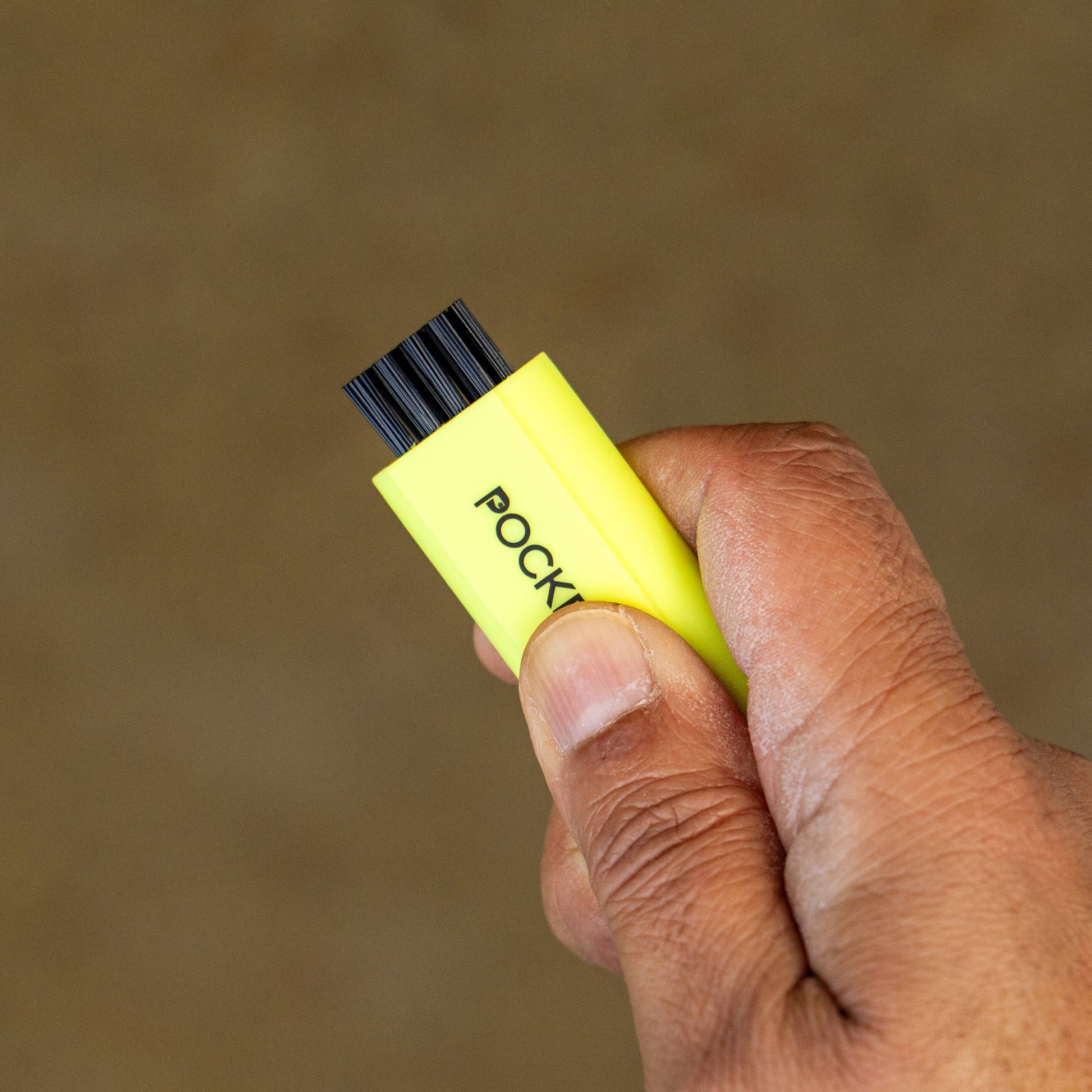 A neon yellow Pocket Brush is being held up with the nylon bristles exposed agains a sandy background.