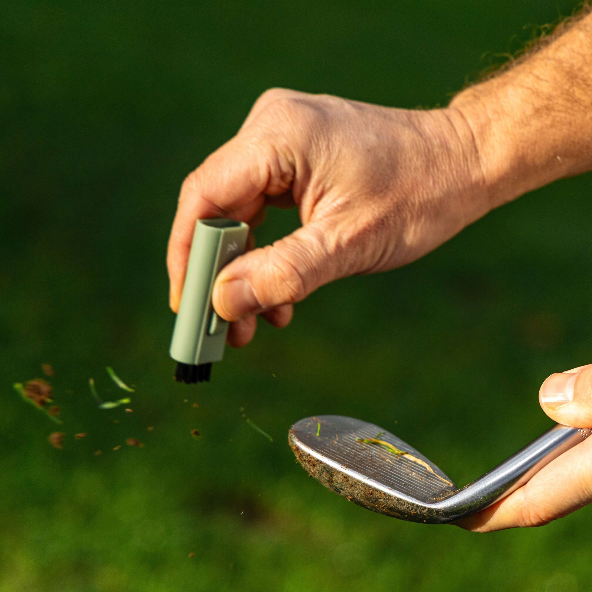 A sage green Pocket Brush is using the nylon bristles to remove debris from a golf club face. The debris is flying towards the left  side of the screen.
