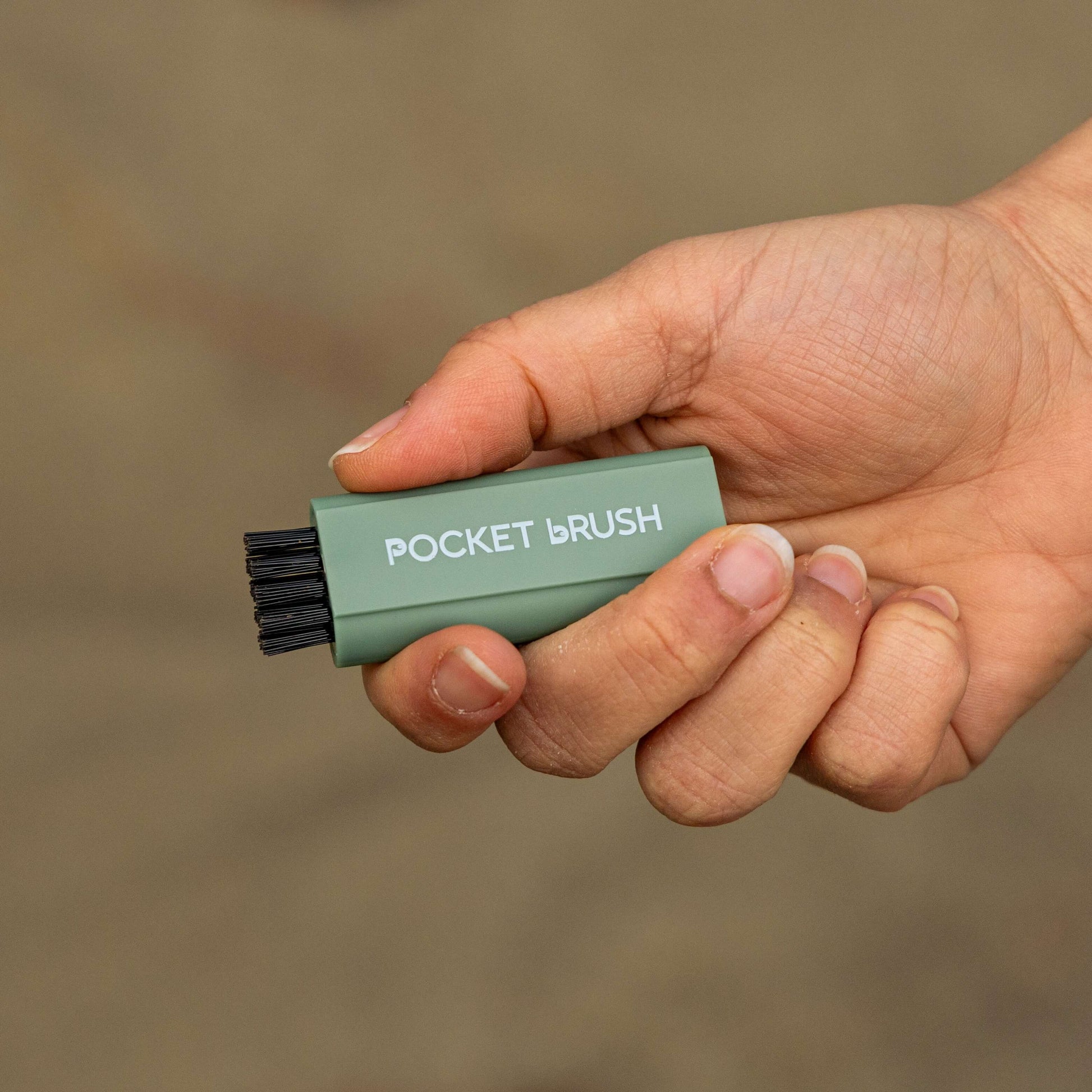 A sage green Pocket Brush golf club brush is being held up with the nylon bristles exposed against a sandy background
