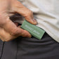 A sage green Pocket Brush is being held next to a black pant pocket.