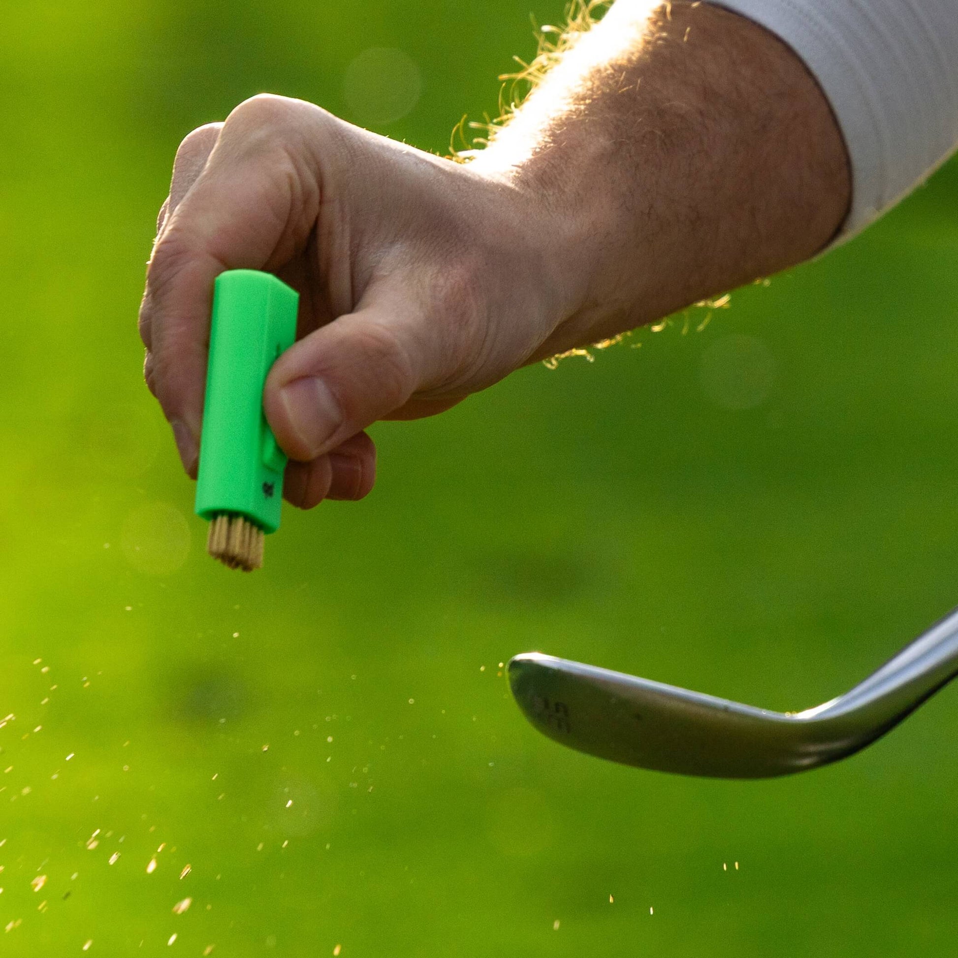A neon green Pocket Brush is using its brass bristles to remove the dirt from a golf club