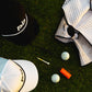 Golf accessories including a pocket brush cap, towel, golf balls, and club on grass