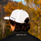 Person wearing a white Pocket Brush cap and black jacket with text, standing in a forest with autumn foliage.