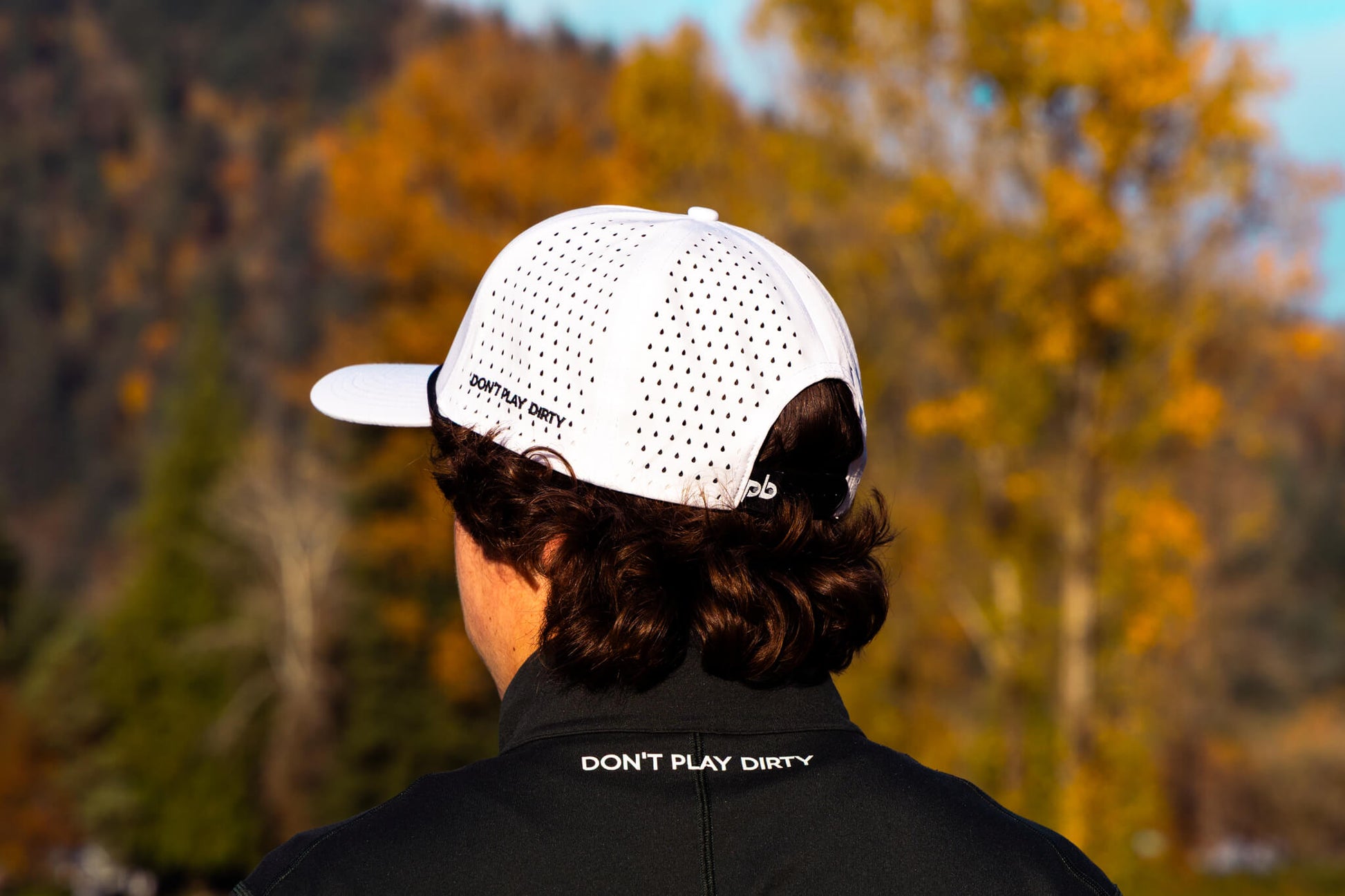 Person wearing a white Pocket Brush cap and black jacket with text, standing in a forest with autumn foliage.