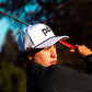 Golfer in action with a golf club, wearing a white cap with a Pocket Brush logo, against a blurred natural background.