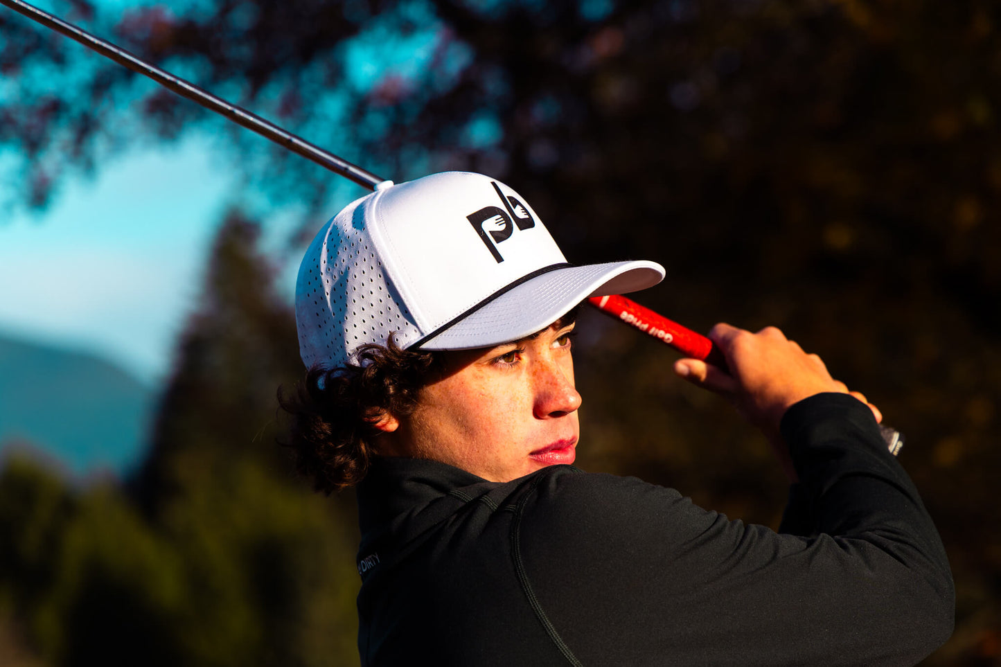 Golfer in action with a golf club, wearing a white cap with a Pocket Brush logo, against a blurred natural background.