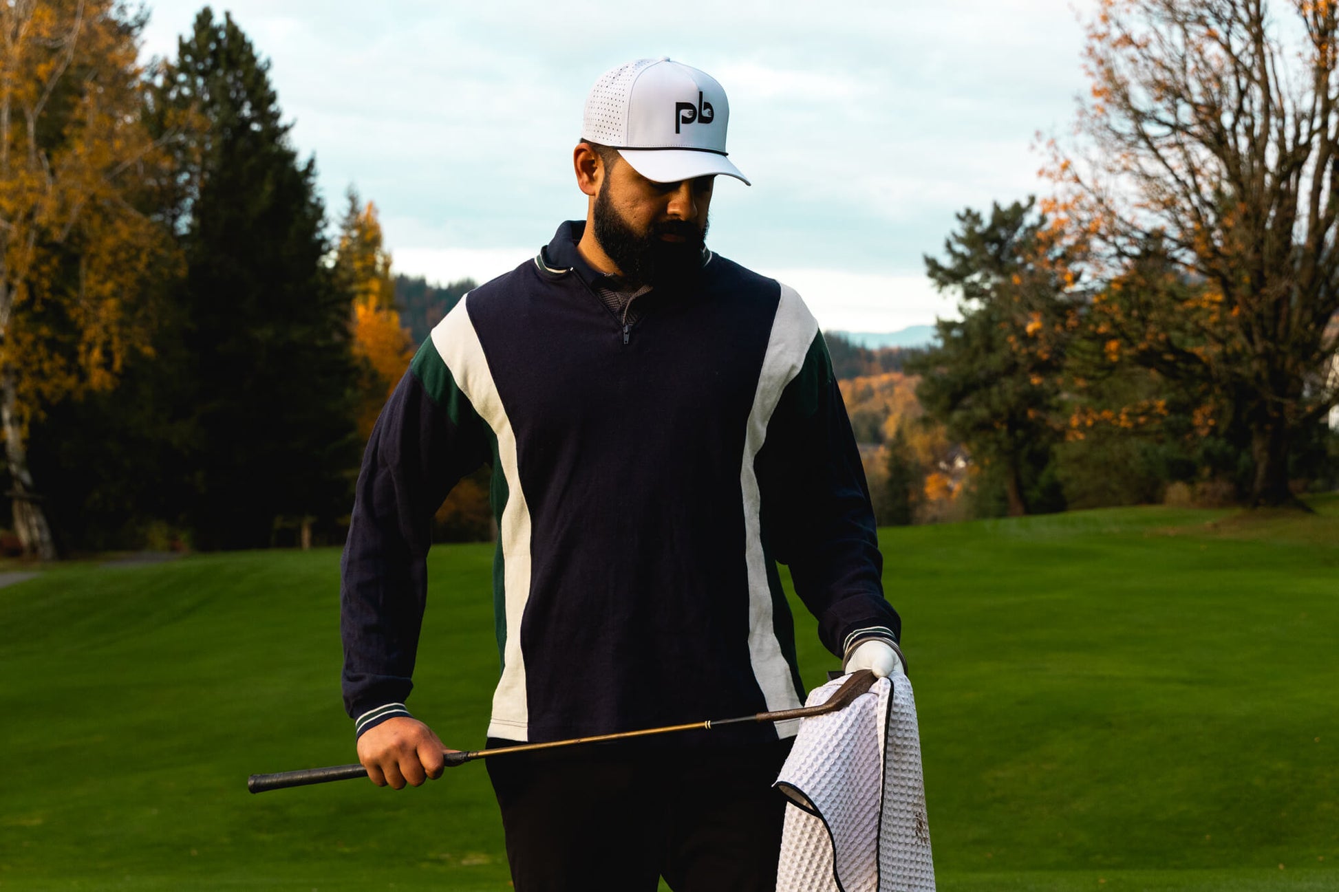 Man with a white pocket brush hat on a golf course holding a golf club with a blurred autumn background
