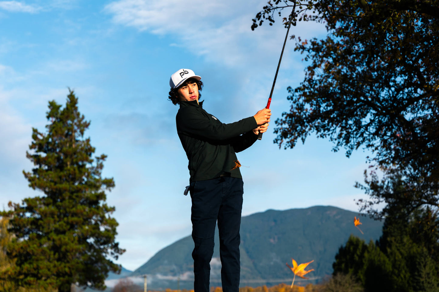 Person playing golf wearing a white pocket brush cap with mountains and trees in the background